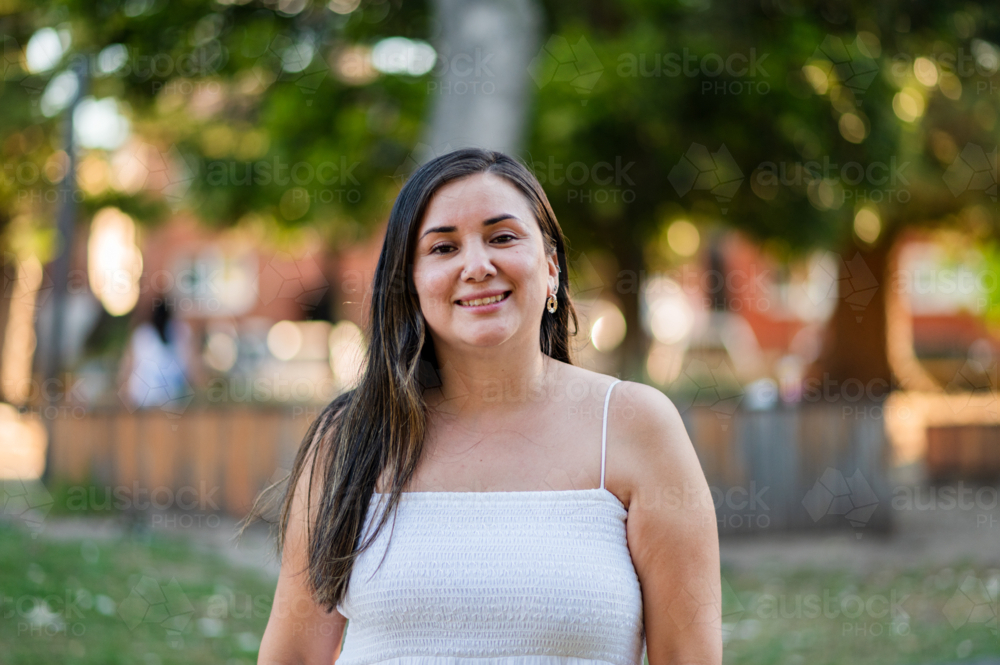 A woman stands in a park smiling while sunlight filters through the trees in the background - Australian Stock Image