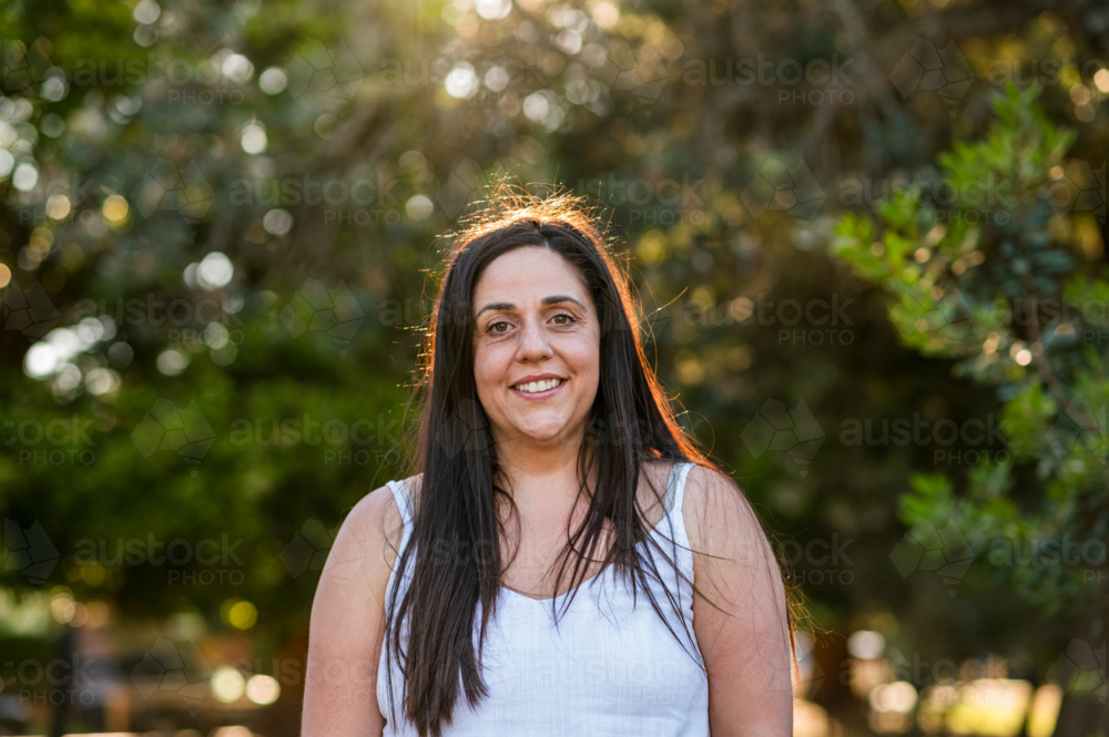 A woman stands in a park surrounded by trees while the sun sets in the background - Australian Stock Image