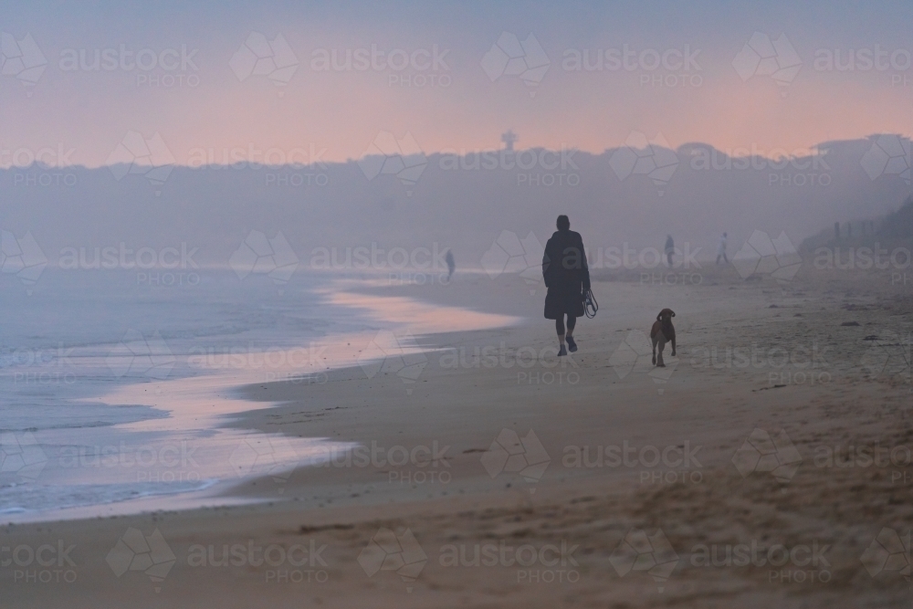 A woman walking with her dog on a foggy beach at sunset - Australian Stock Image