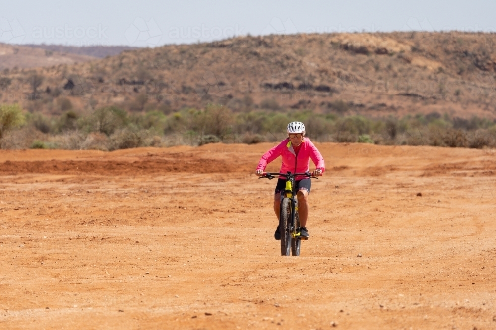 A woman wearing bright pink top riding a mountain bike through an outback landscape - Australian Stock Image
