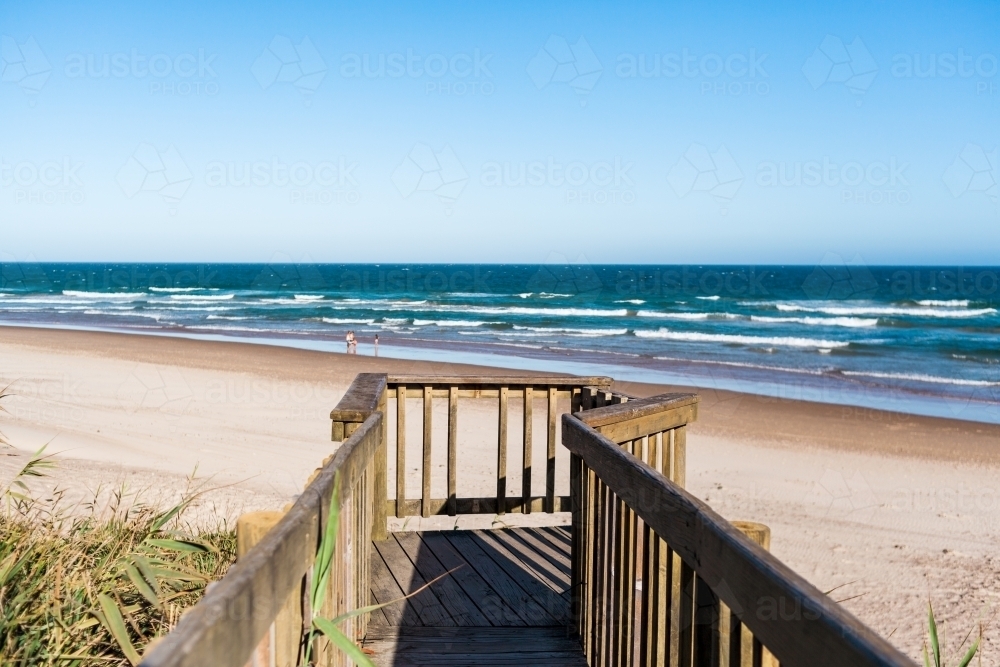 A wooden, narrow viewing platform looks out over a people enjoying a sunny day at the beach. - Australian Stock Image
