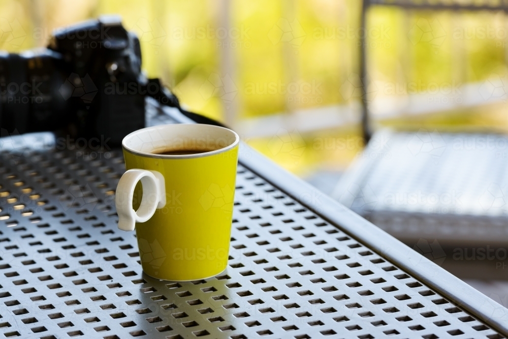A yellow mug of coffee on a metal outdoor table with blurred camera and chair in background - Australian Stock Image