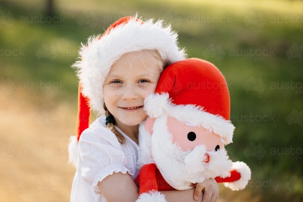 A young australian caucasian girl wearing a Christmas hat and cuddling a stuffed Santa clause toy - Australian Stock Image
