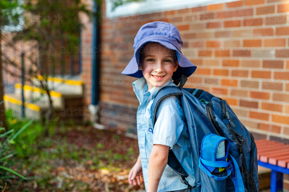 A young boy in a school uniform stands outside a primary school wearing a hat and smiling - Australian Stock Image