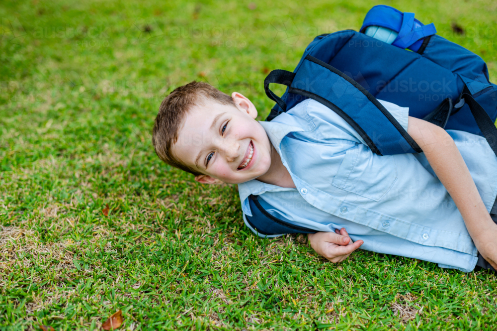 A young boy in school uniform is lying on the grass with a backpack while smiling - Australian Stock Image