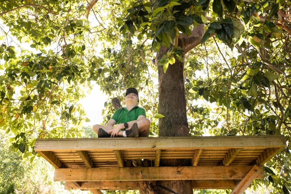 A young boy laughing while wearing a hat sitting in a backward tree house in an australian backyard - Australian Stock Image