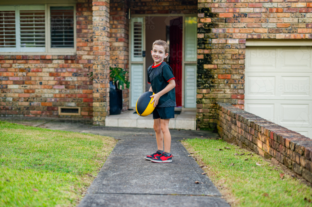 A young boy plays on the pathway outside a brick house while holding a rugby ball and smiling - Australian Stock Image