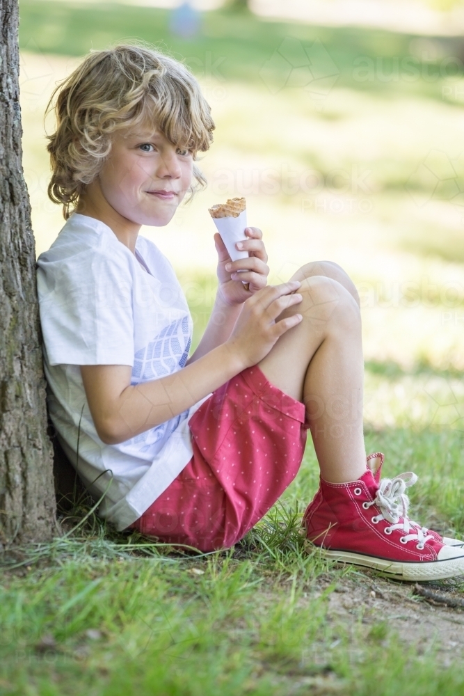 A young boy sitting against a tree eating an ice cream and smiling - Australian Stock Image