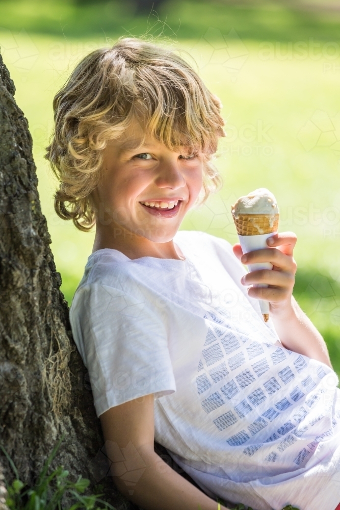 A young boy sitting against a tree eating an ice cream cone - Australian Stock Image
