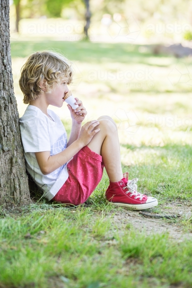 A young boy sitting against a tree eating an icecream cone - Australian Stock Image