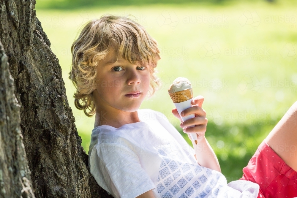A young boy sitting against a tree holding an ice cream - Australian Stock Image