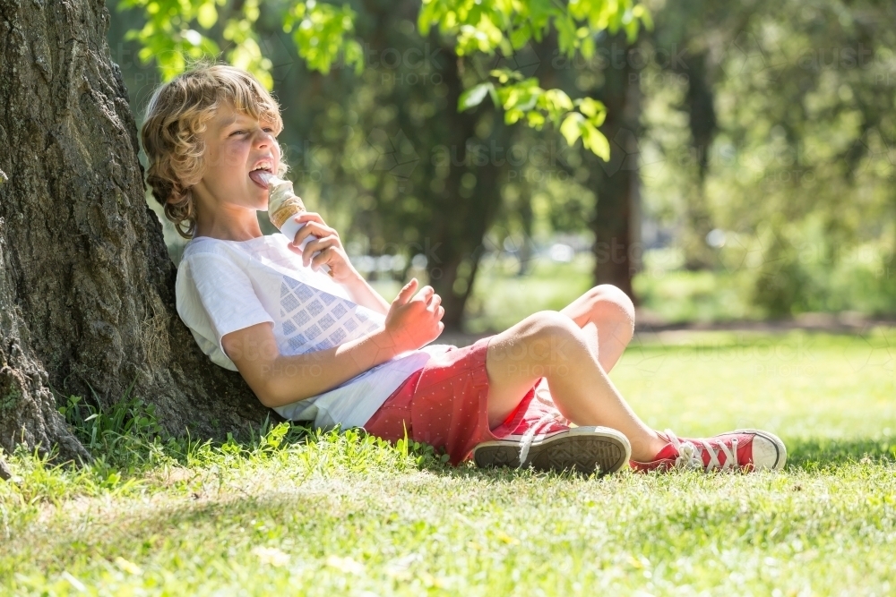 A young boy sitting against a tree licking an ice cream - Australian Stock Image