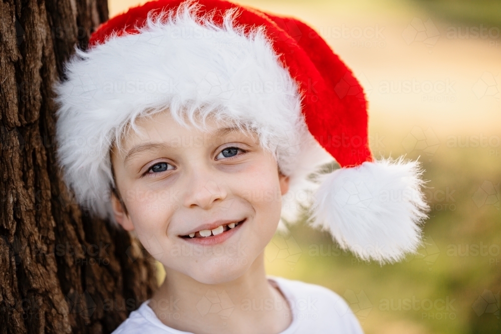 A young caucasian Australian boy wearing a Santa hat leaning against a tree and smiling at Christmas - Australian Stock Image