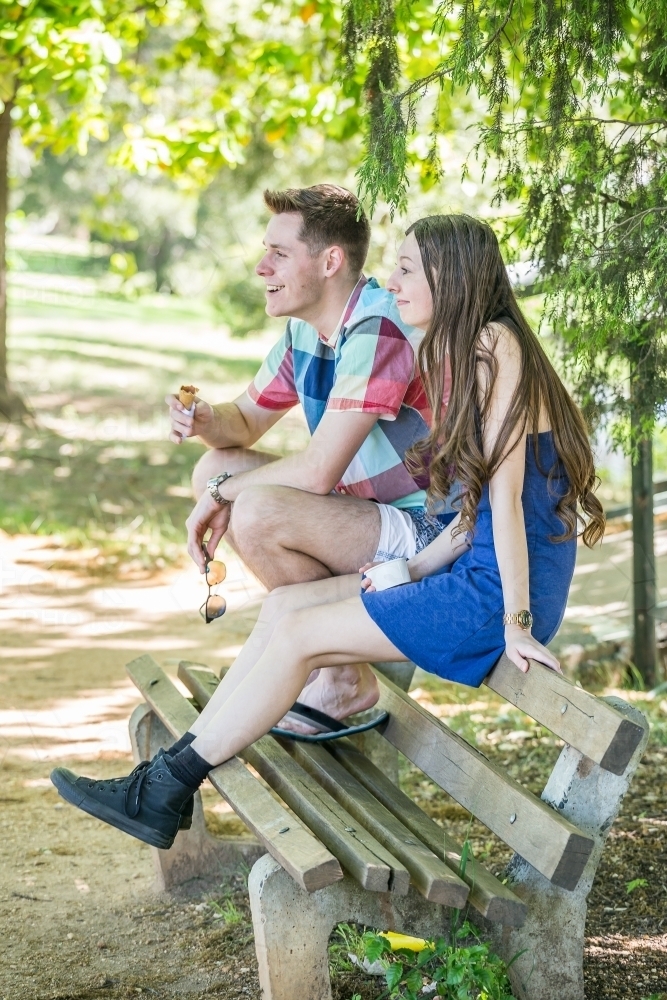 A young couple sit on a park bench eating ice cream - Australian Stock Image