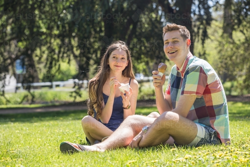 A young couple sitting on the ground eating icecreams in a park - Australian Stock Image