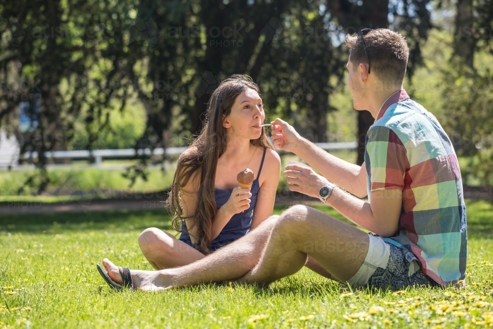 A young couple sitting on the ground sharing ice creams in a park - Australian Stock Image
