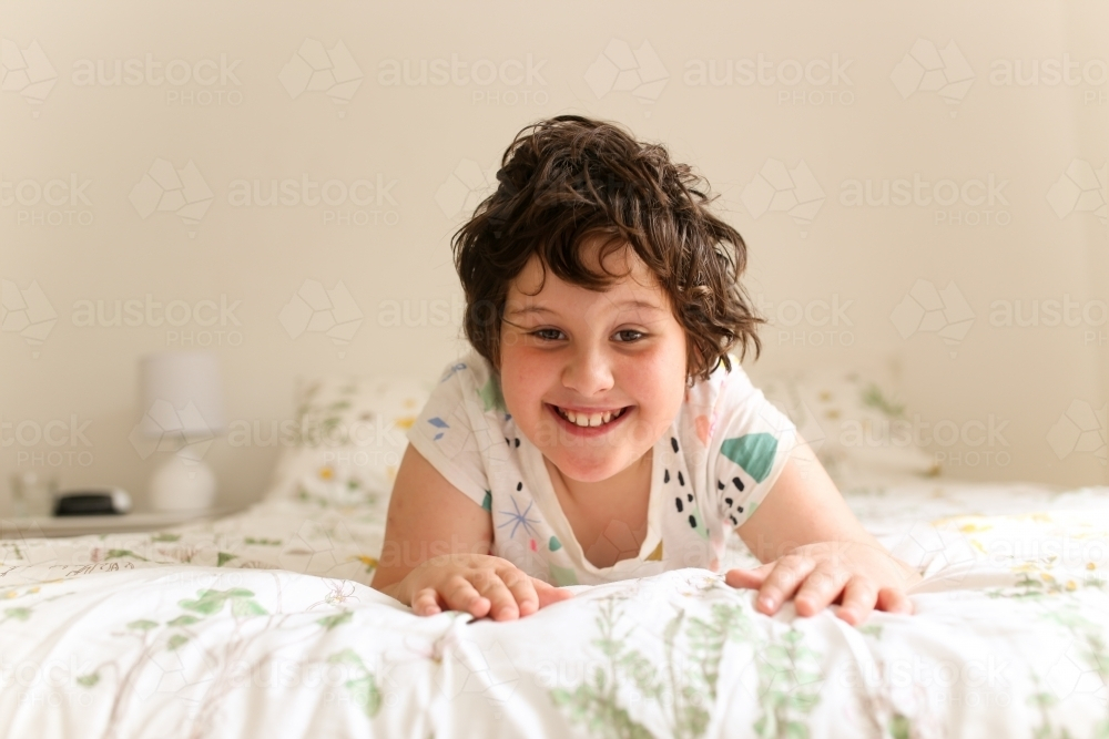 A young girl lying on her tummy on a bed smiling - Australian Stock Image