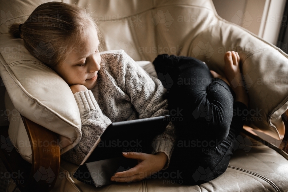 A young girl of caucasion decent sitting by a window watching programs on her tablet - Australian Stock Image