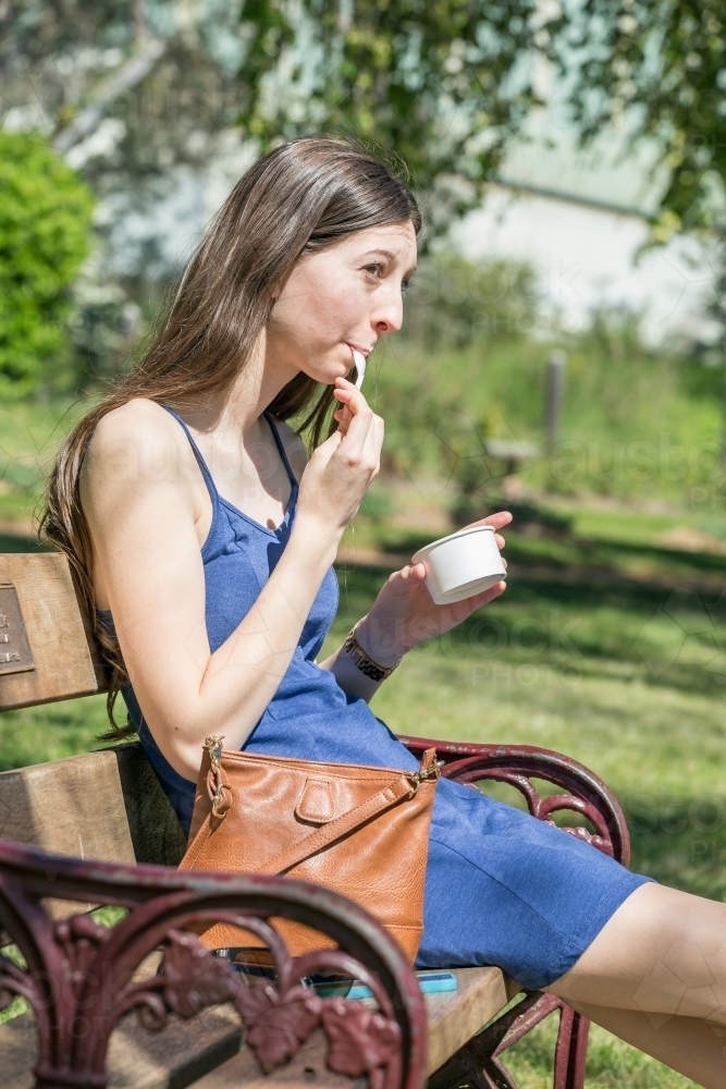 A young lady sits on a park bench eating icecream out of a cup - Australian Stock Image