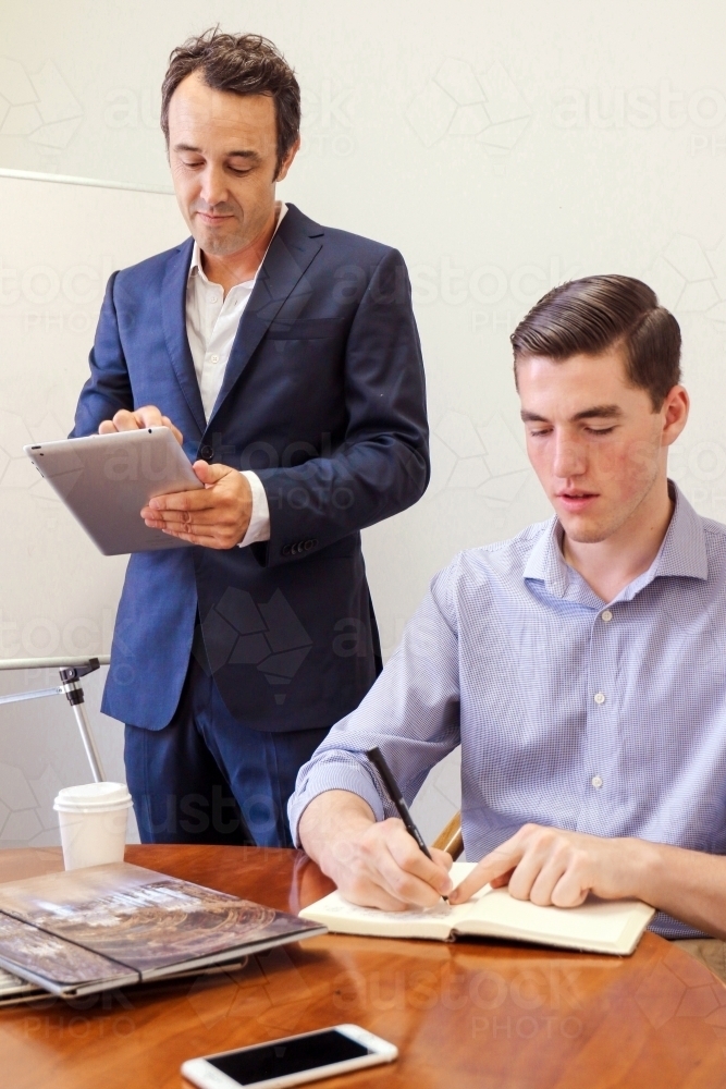 A young male office worker taking notes at a meeting desk with manager standing behind - Australian Stock Image