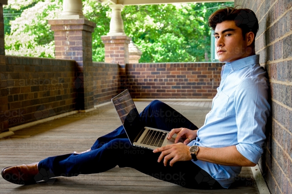 A young male sitting on the ground on a covered verandah with a laptop - Australian Stock Image