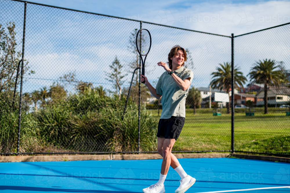 A young male tennis player smiles on a blue court amid greenery and palm trees - Australian Stock Image