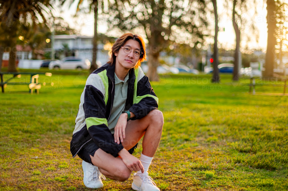 A young man crouches on the grass wearing casual sports clothes as the sun sets nearby - Australian Stock Image
