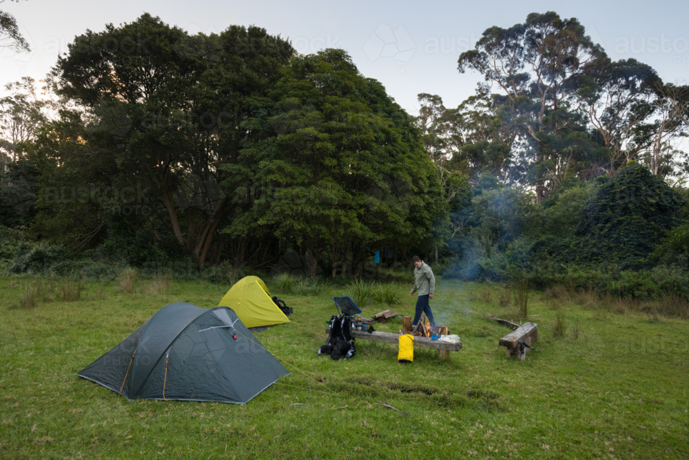 A young man in his twenties walks next to a campfire with small tents nearby - Australian Stock Image