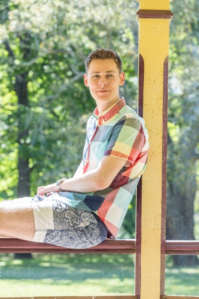 A young man sitting relaxed on a wooden railing in a park - Australian Stock Image