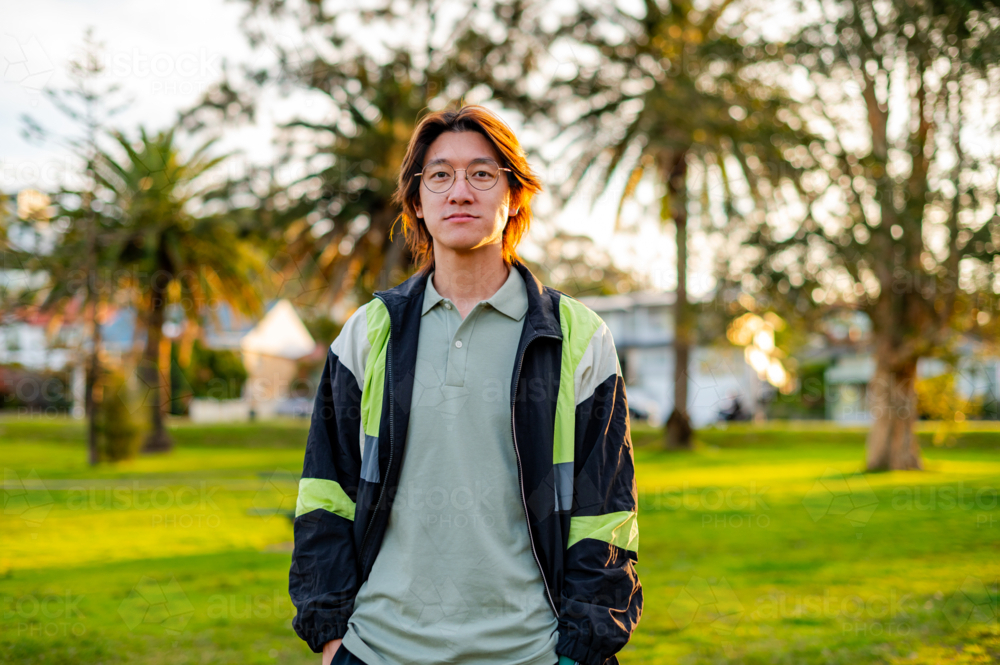 A young man wearing glasses and a light green top poses in a park surrounded by palm trees - Australian Stock Image
