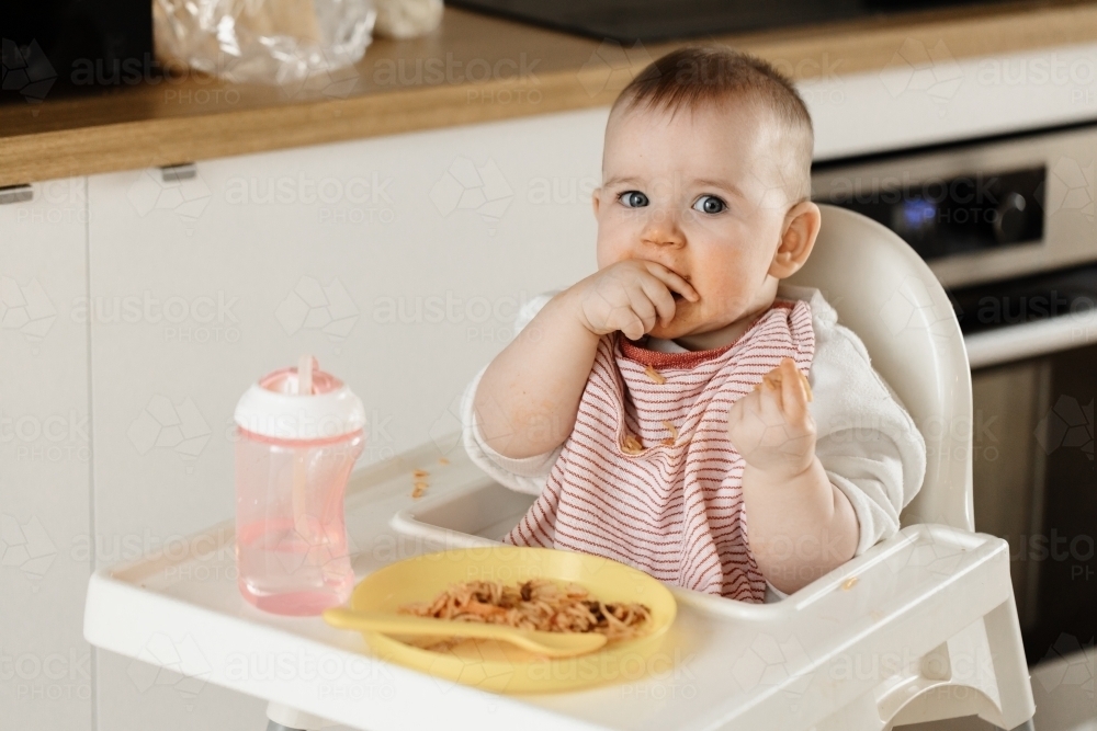 Image of A young one year old girl child sitting in a high chair eating
