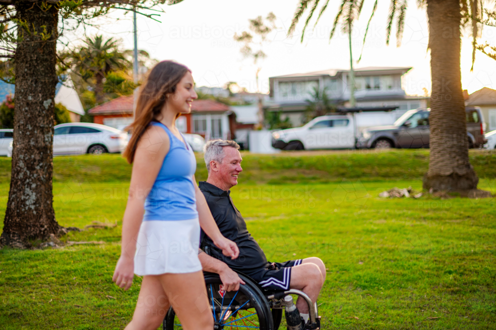 A young woman walks alongside a man in a wheelchair at a park, enjoying sunset together - Australian Stock Image