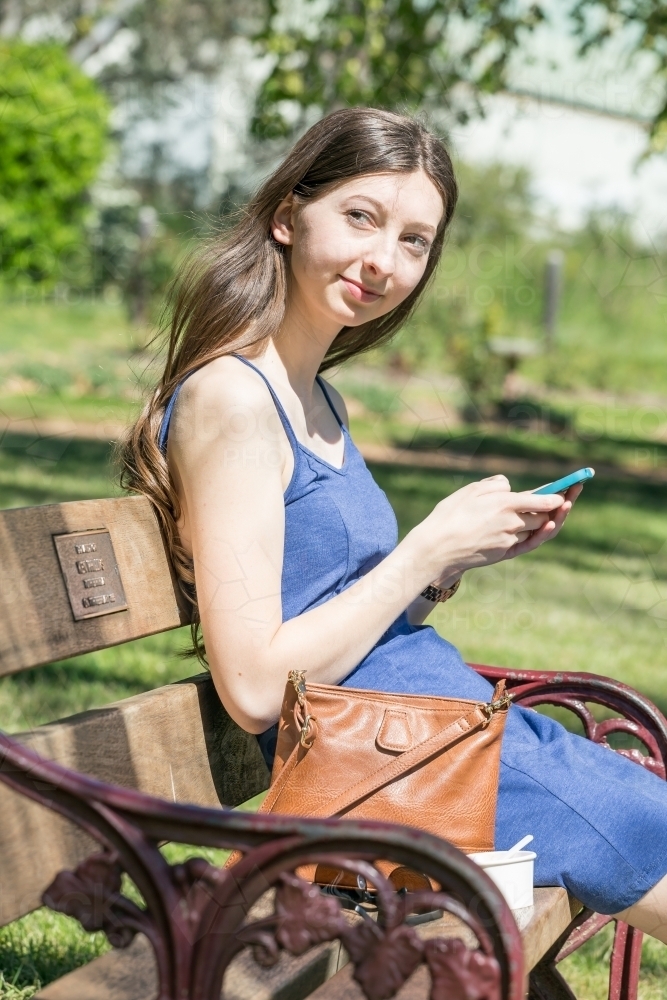 A young woman with long hair sitting on a park bench with her phone in her hands - Australian Stock Image