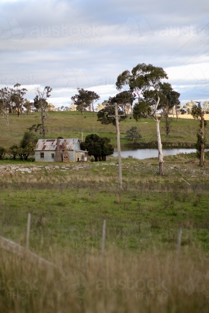 Abandoned work mans hut situated in a paddock by a dam full of water in the afternoon - Australian Stock Image