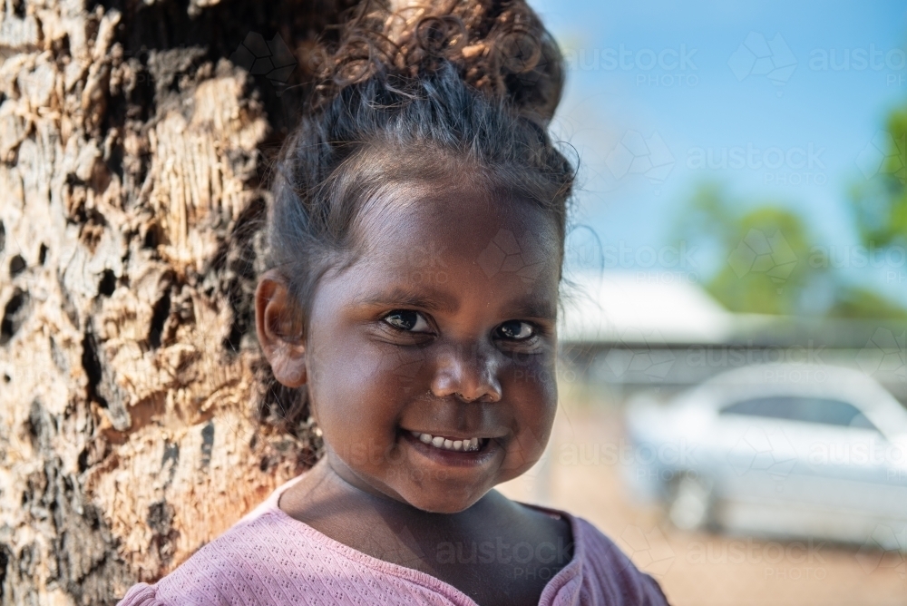 Aboriginal 3 year old girl smiling by a tree - Australian Stock Image