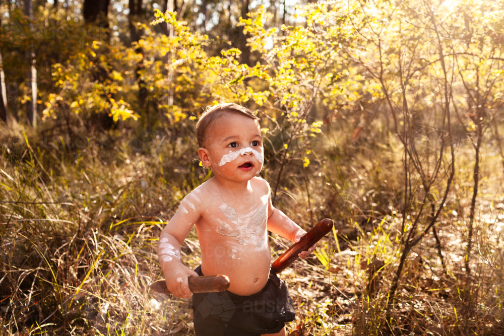 Aboriginal Australian toddler boy painted in ochre in green natural bushland with clapsticks - Australian Stock Image