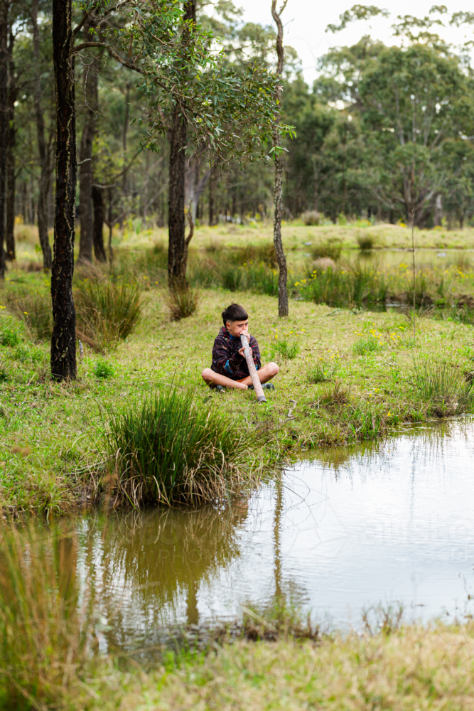 aboriginal boy playing didgeridoo by creek in green bushland - Australian Stock Image