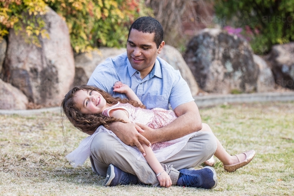 Aboriginal father sitting on ground smiling down at mixed race daughter lying in arms - Australian Stock Image