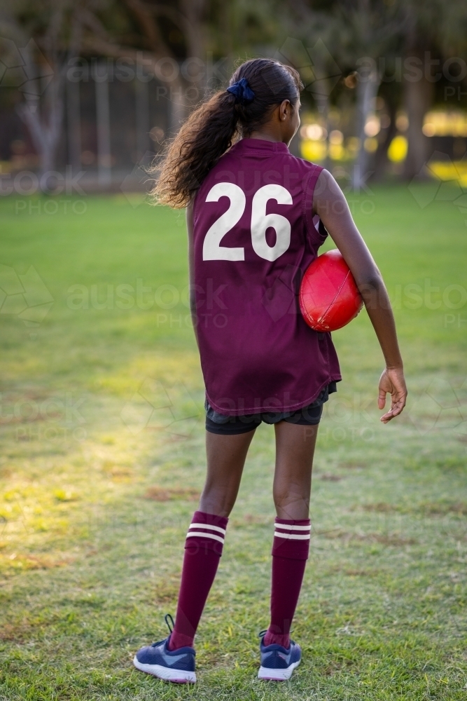 aboriginal girl in football uniform holding ball under her arm seen from behind - Australian Stock Image