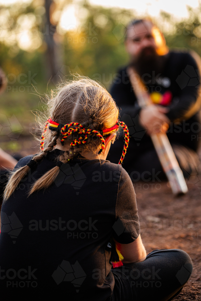 Aboriginal girl listening to man playing didgeridoo in fading light in Australian bushland - Australian Stock Image