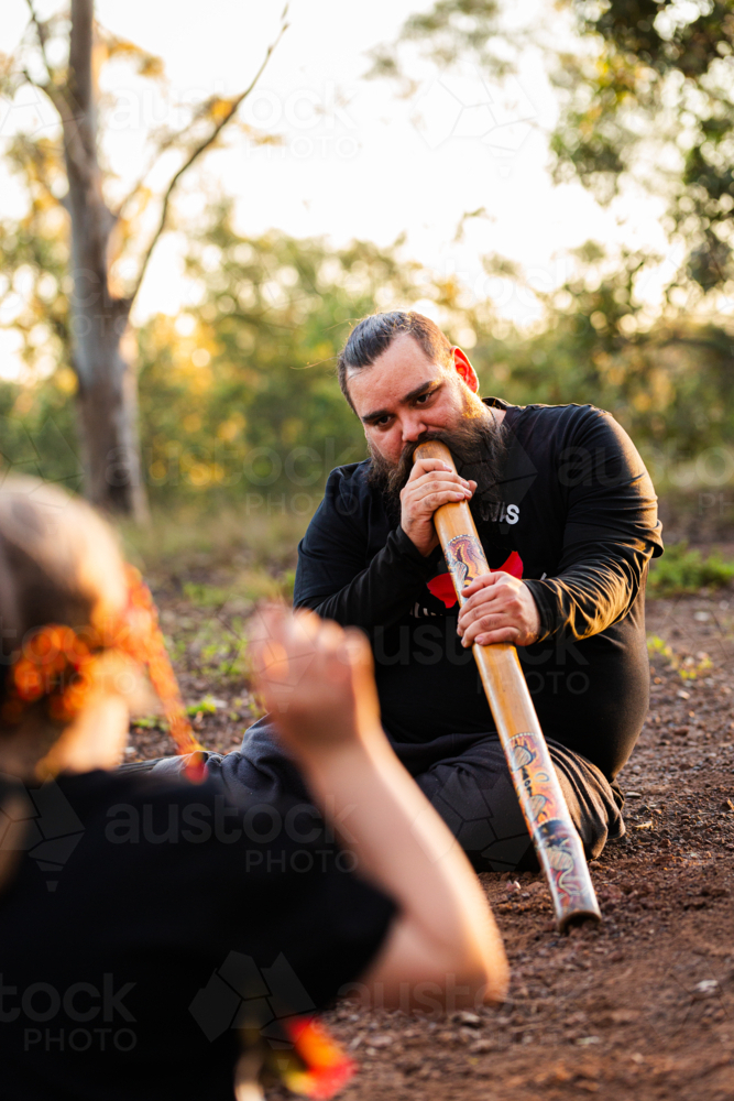 Aboriginal girl listening to man playing didgeridoo in golden light in Australian bushland - Australian Stock Image