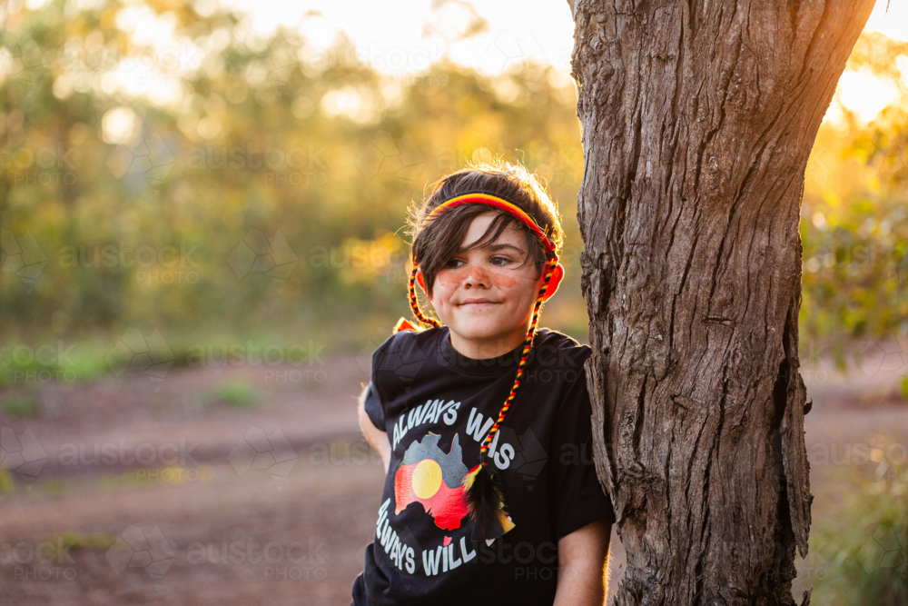 Aboriginal kid with red yellow and black headband leaning on tree at sunset - Australian Stock Image