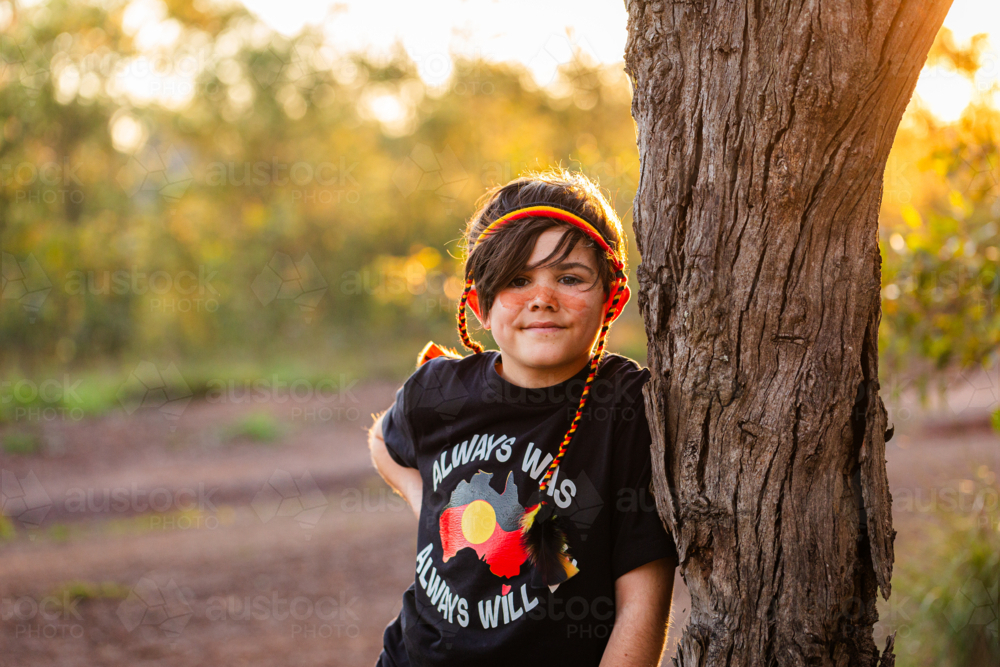 Aboriginal kid with red yellow and black headband leaning on tree at sunset - Australian Stock Image