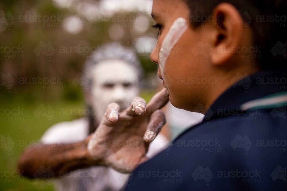 Aboriginal man wearing white face paint putting white face paint onto a young Aboriginal boy - Australian Stock Image