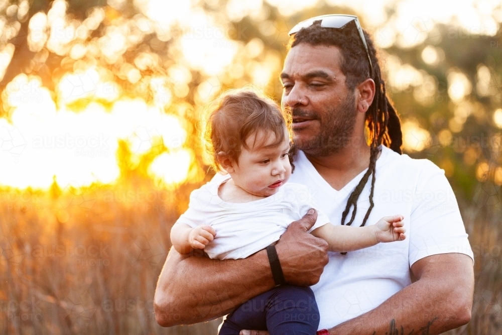 Aboriginal man with baby daughter in country setting at sunset - Australian Stock Image
