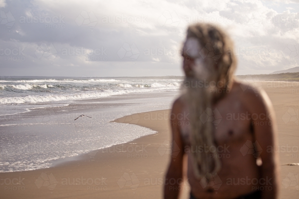 Aboriginal man with clay face paint standing on a beach focus on the water and sand - Australian Stock Image