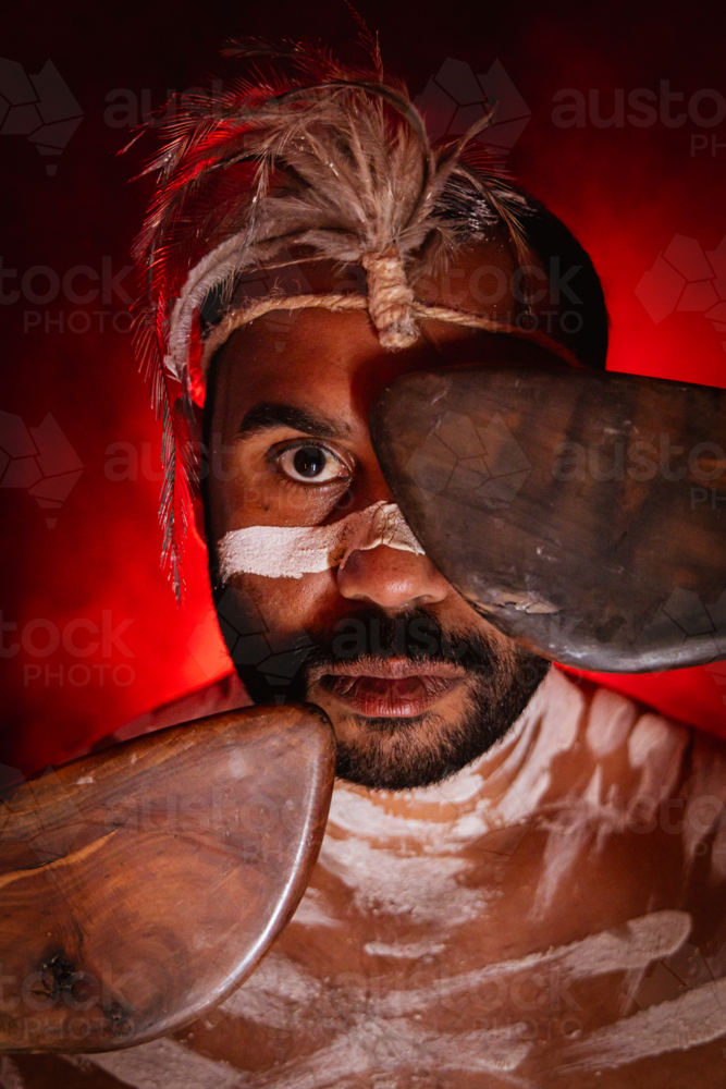 Aboriginal man with red background covering one eye with boomerang - Australian Stock Image