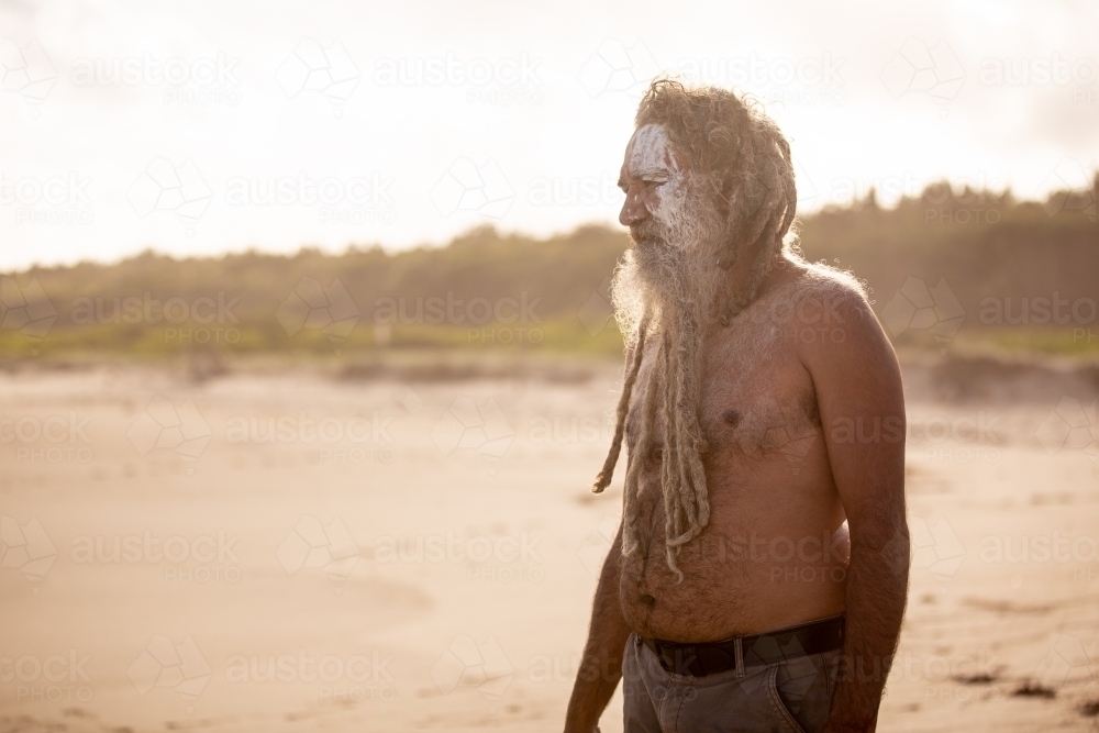 Aboriginal middle aged man with clay face paint standing on a beach - Australian Stock Image