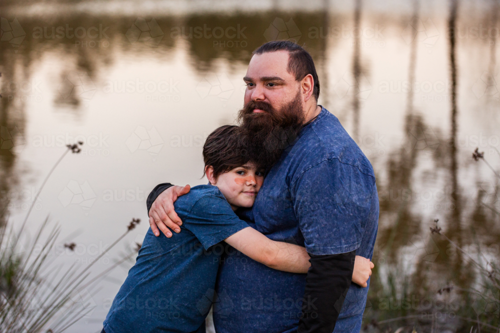 Aboriginal son hugging father outdoors by waterside at the end of the day - Australian Stock Image