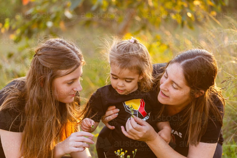 Aboriginal toddler playing outside in nature with big sister and mum looking at flowers - Australian Stock Image